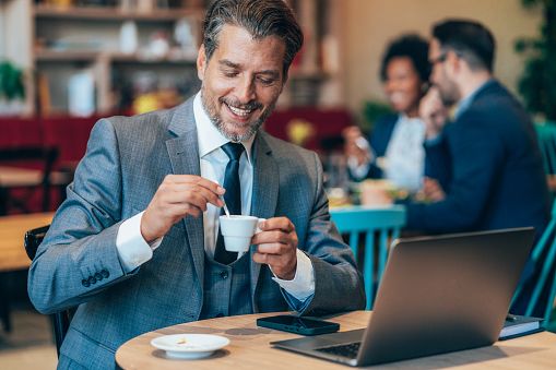 Lächelnder Mann um die vierzig in einem Café mit Laptop und einer Tasse Kaffee, der eine ernsthafte Begegnung, einen positiven ersten Eindruck und authentische Kommunikation vermittelt.