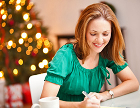 Single_Frau sitzt an Schreibtisch und schreibt einen Brief. Hinter ihr steht ein Weihnachtsbaum